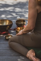 A person sits cross-legged on a wooden floor, surrounded by several singing bowls and a mallet. The person is dressed in brown leggings and a sleeveless top. The scene is bathed in dappled natural light, creating a serene and meditative atmosphere.