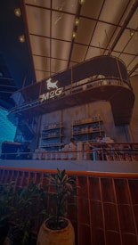 An interior setting of a modern caf&eacute; or bar with a stylish, elevated seating area labeled 'MoG' on a sign featuring a dog graphic. There are plants in the foreground, and shelves stocked with bottles against the wall. The ceiling is adorned with hanging lights, giving the space a warm ambiance. Two staff members are visible behind the counter.