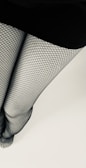 A pair of legs is covered with fishnet stockings, standing against a light background. The skirt or dress at the top is black, contrasting with the patterned texture of the stockings.