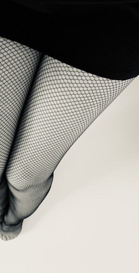 A pair of legs is covered with fishnet stockings, standing against a light background. The skirt or dress at the top is black, contrasting with the patterned texture of the stockings.