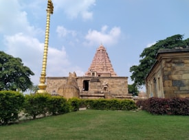 A historical temple structure made of stone with intricate carvings can be seen, surrounded by neatly maintained lawns and greenery. A tall golden pole stands prominently to the left, and scattered clouds decorate the blue sky above.