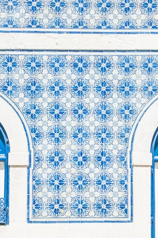 A wall adorned with intricate blue and white tiles forming an elaborate geometric pattern. The tiles are organized in symmetrical floral motifs, bordered by white stone with an arched design. The sunlight casts a bright illumination on the surface, highlighting the vivid blue and delicate detailing of each tile.
