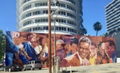 A vibrant mural depicting several jazz musicians and prominent figures is painted on a wall in front of a modern, circular building. The mural includes people playing instruments like trumpets and saxophones, and shows a diverse group of individuals, some smiling warmly. The scene is set under a clear blue sky, with palm trees and modern architecture in the background.