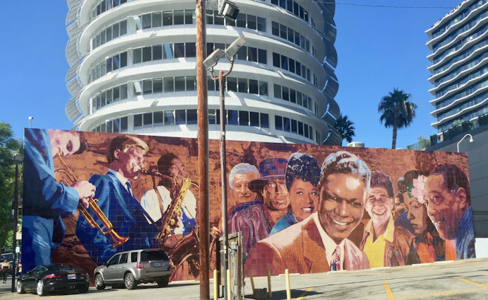 A vibrant mural depicting several jazz musicians and prominent figures is painted on a wall in front of a modern, circular building. The mural includes people playing instruments like trumpets and saxophones, and shows a diverse group of individuals, some smiling warmly. The scene is set under a clear blue sky, with palm trees and modern architecture in the background.