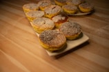 A collection of small, round pastries topped with powdered sugar arranged on a rectangular white plate and a round plate in the background. The pastries are placed on a wooden table lightly dusted with flour, creating a warm and inviting atmosphere.