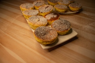 A collection of small, round pastries topped with powdered sugar arranged on a rectangular white plate and a round plate in the background. The pastries are placed on a wooden table lightly dusted with flour, creating a warm and inviting atmosphere.