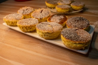 A wooden table displays a white rectangular platter with several freshly baked pastries. They are round with a slightly cracked top and dusted with powdered sugar. In the background, there is a plate with more pastries slightly out of focus.