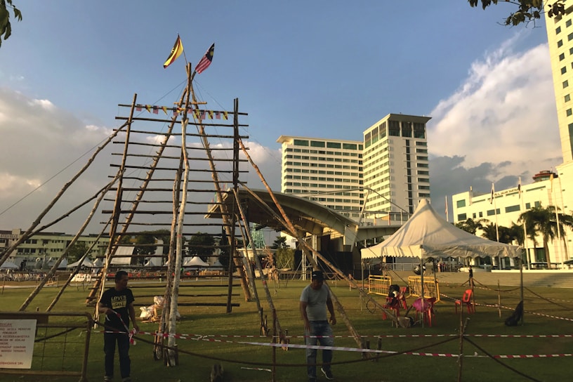 A large wooden structure resembling a traditional bamboo swing stands prominently in an open grassy area. Several people are present, setting up or engaging with the structure. Flags are mounted at the top of the swing. In the background, modern buildings and a white tent are visible under a partly cloudy sky.