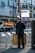 A man in a dark suit is standing in front of a building at an intersection, examining a signboard. The urban environment features prominent signage, a truck with an orange cover parked on the street, and modern architecture. The atmosphere suggests a busy city scene, possibly in a commercial district.