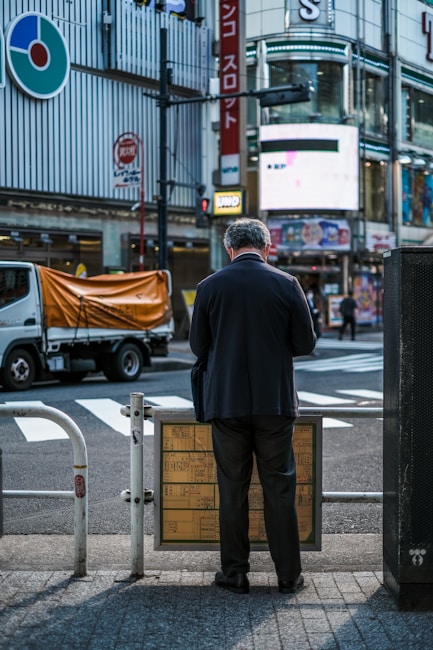 A man in a dark suit is standing in front of a building at an intersection, examining a signboard. The urban environment features prominent signage, a truck with an orange cover parked on the street, and modern architecture. The atmosphere suggests a busy city scene, possibly in a commercial district.