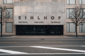 A modern, minimalist building facade with a sign that reads 'SIHLHOF' in large letters. The building features a concrete front and large rectangular windows. Two bare trees are situated along the sidewalk flanking the entrance, which has dark tinted glass doors. The street in front has clearly marked lanes and arrows.