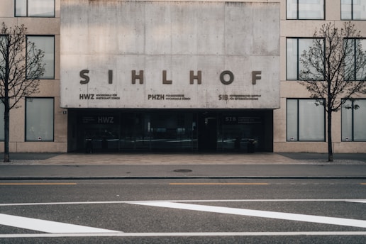 A modern, minimalist building facade with a sign that reads 'SIHLHOF' in large letters. The building features a concrete front and large rectangular windows. Two bare trees are situated along the sidewalk flanking the entrance, which has dark tinted glass doors. The street in front has clearly marked lanes and arrows.