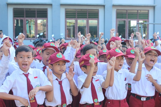 A group of young children wearing red and white uniforms with matching hats are energetically raising their fists in the air. They appear to be outside a building with multiple large windows. The children are of similar age and all seem to be engaged in some form of group activity.
