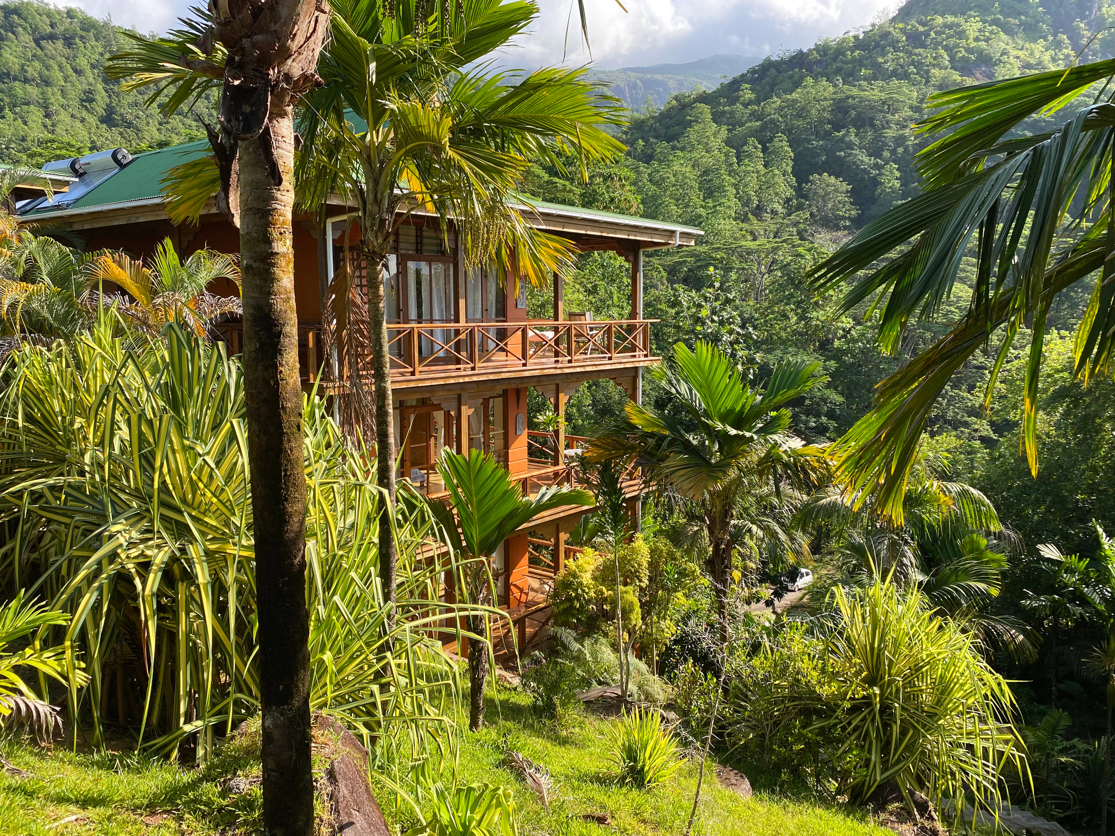 A three-storey timber guesthouse with open balconies and a green roof stands among coconut palms and screwpine on a steep forested hillside on the west coast of Mahe