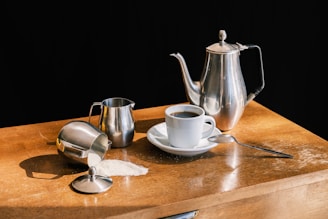 On a wooden surface, there is a set of metal kitchenware: a tall teapot, a small milk jug, and a sugar bowl with some sugar spilled nearby. A white coffee cup filled with black coffee is placed on a saucer next to a small spoon.