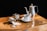 On a wooden surface, there is a set of metal kitchenware: a tall teapot, a small milk jug, and a sugar bowl with some sugar spilled nearby. A white coffee cup filled with black coffee is placed on a saucer next to a small spoon.