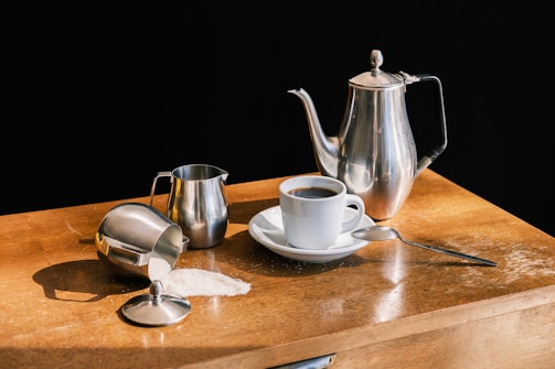 On a wooden surface, there is a set of metal kitchenware: a tall teapot, a small milk jug, and a sugar bowl with some sugar spilled nearby. A white coffee cup filled with black coffee is placed on a saucer next to a small spoon.