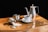 On a wooden surface, there is a set of metal kitchenware: a tall teapot, a small milk jug, and a sugar bowl with some sugar spilled nearby. A white coffee cup filled with black coffee is placed on a saucer next to a small spoon.