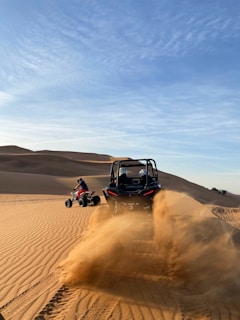 Two off-road vehicles are being driven through a sandy desert. The vehicle in the foreground is on the right side, kicking up a large cloud of sand, while the vehicle on the left side is smaller in the distance with a person riding an all-terrain vehicle (ATV). The landscape is characterized by sand dunes under a clear blue sky with wisps of clouds.