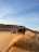 Two off-road vehicles are being driven through a sandy desert. The vehicle in the foreground is on the right side, kicking up a large cloud of sand, while the vehicle on the left side is smaller in the distance with a person riding an all-terrain vehicle (ATV). The landscape is characterized by sand dunes under a clear blue sky with wisps of clouds.