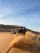 Two off-road vehicles are being driven through a sandy desert. The vehicle in the foreground is on the right side, kicking up a large cloud of sand, while the vehicle on the left side is smaller in the distance with a person riding an all-terrain vehicle (ATV). The landscape is characterized by sand dunes under a clear blue sky with wisps of clouds.