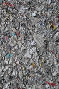 A chaotic mix of shredded paper, displaying various colors and text fragments. The paper pieces are in different sizes and orientations, creating a textured, layered appearance. Bits of text in various languages are discernible among the fragments, alongside a predominance of gray, white, and muted colors.