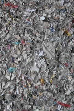 A chaotic mix of shredded paper, displaying various colors and text fragments. The paper pieces are in different sizes and orientations, creating a textured, layered appearance. Bits of text in various languages are discernible among the fragments, alongside a predominance of gray, white, and muted colors.