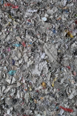 A chaotic mix of shredded paper, displaying various colors and text fragments. The paper pieces are in different sizes and orientations, creating a textured, layered appearance. Bits of text in various languages are discernible among the fragments, alongside a predominance of gray, white, and muted colors.