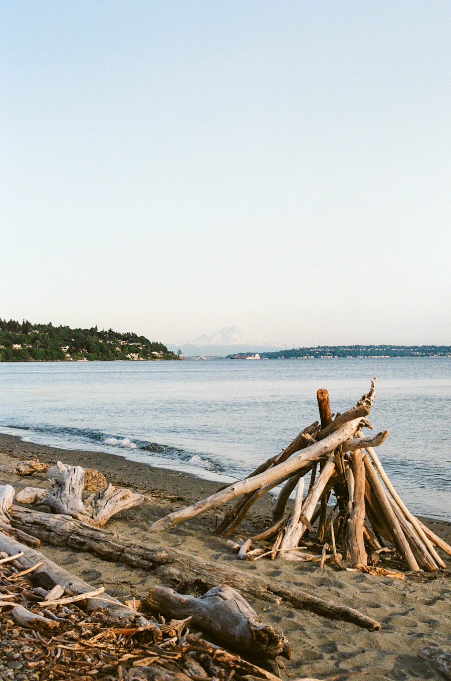 A driftwood-strewn Puget Sound beach near Burien Washington at golden hour, with a snow-capped peak across the Sound