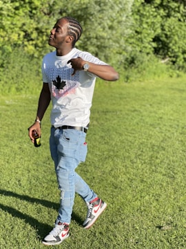 A person with braided hair stands on a grassy field holding a bottle. They are wearing a white T-shirt with a printed design, light blue jeans, and sneakers. The background is filled with lush green foliage, suggesting a park or natural setting.