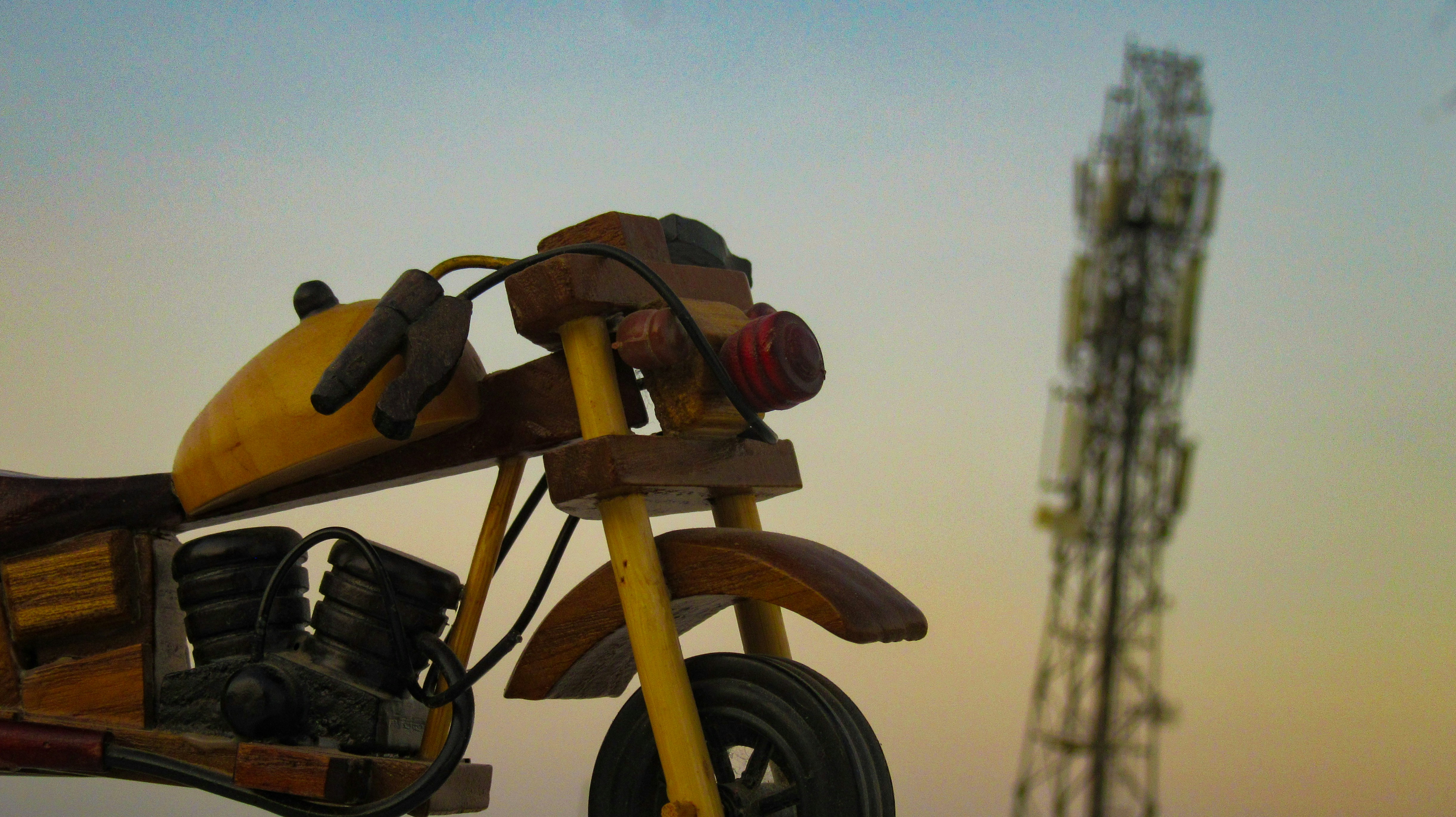 Close-up of a wooden toy motorcycle silhouetted against a warm sunset, with a distant cell tower in the background.