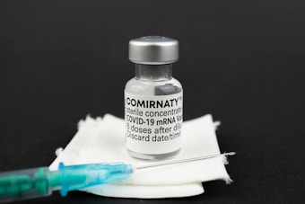 A vial labeled 'Comirnaty' containing COVID-19 mRNA vaccine is positioned on a white pad. A syringe with a teal-colored needle is placed beside the vial. The background is black, emphasizing the medical items.