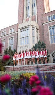 A group of young women in white dresses with red sashes stand in a line in front of a large, ornate brick building with tall windows and detailed architecture. The foreground features colorful flowers and greenery, adding a vibrant touch to the scene.