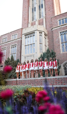 A group of young women in white dresses with red sashes stand in a line in front of a large, ornate brick building with tall windows and detailed architecture. The foreground features colorful flowers and greenery, adding a vibrant touch to the scene.