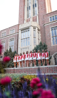A group of young women in white dresses with red sashes stand in a line in front of a large, ornate brick building with tall windows and detailed architecture. The foreground features colorful flowers and greenery, adding a vibrant touch to the scene.