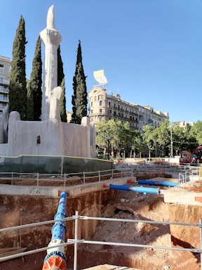A construction site in an urban environment features a deep excavation with visible pipes and a fenced-off area. Large cypress trees and a covered monument or sculpture are present in the foreground. In the background, there are historic buildings with ornamental facades and a prominent owl sign atop one of the buildings. The sky is clear and blue, providing a contrast to the earthy tones of the construction site.