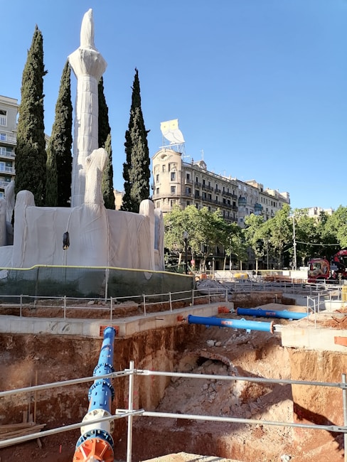 A construction site in an urban environment features a deep excavation with visible pipes and a fenced-off area. Large cypress trees and a covered monument or sculpture are present in the foreground. In the background, there are historic buildings with ornamental facades and a prominent owl sign atop one of the buildings. The sky is clear and blue, providing a contrast to the earthy tones of the construction site.