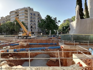 An urban construction site with a large crane and deep excavation. The site is surrounded by buildings and trees, with protective barriers and construction equipment visible.