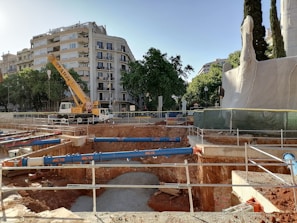 An urban construction site with a large crane and deep excavation. The site is surrounded by buildings and trees, with protective barriers and construction equipment visible.
