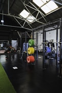 A spacious gym interior with high ceilings and large skylights providing natural light. Various gym equipment, including weight racks with colorful plates (green, yellow, red, blue), a pull-up rig, and exercise machines are neatly organized. The space has a mix of black flooring and a strip of green turf, creating a clean and organized workout environment.