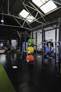 A spacious gym interior with high ceilings and large skylights providing natural light. Various gym equipment, including weight racks with colorful plates (green, yellow, red, blue), a pull-up rig, and exercise machines are neatly organized. The space has a mix of black flooring and a strip of green turf, creating a clean and organized workout environment.