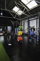 A spacious gym interior with high ceilings and large skylights providing natural light. Various gym equipment, including weight racks with colorful plates (green, yellow, red, blue), a pull-up rig, and exercise machines are neatly organized. The space has a mix of black flooring and a strip of green turf, creating a clean and organized workout environment.