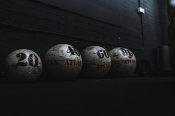 The image depicts four large weighted balls arranged in a row against a dark background. Each ball is labeled with a number indicating its weight, such as 20, 40, 60, and another obscured number. The setting suggests a gym or fitness environment, characterized by dim lighting and a focus on the rugged texture of the equipment.