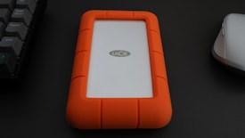 A portable hard drive with an orange rubber casing is placed on a dark surface. To the left is part of a keyboard, and to the right is a white computer mouse.