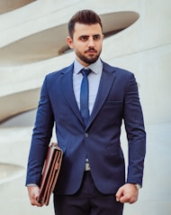 A man in a business suit stands confidently holding a leather briefcase. The background features a modern architectural wall with curved lines. The man has a neatly groomed beard and styled hair, wearing a dark blue suit with a matching tie and a striped shirt.