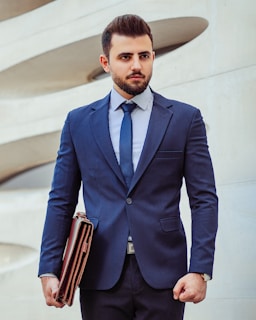 A man in a business suit stands confidently holding a leather briefcase. The background features a modern architectural wall with curved lines. The man has a neatly groomed beard and styled hair, wearing a dark blue suit with a matching tie and a striped shirt.
