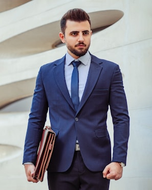 A man in a business suit stands confidently holding a leather briefcase. The background features a modern architectural wall with curved lines. The man has a neatly groomed beard and styled hair, wearing a dark blue suit with a matching tie and a striped shirt.