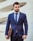A man in a business suit stands confidently holding a leather briefcase. The background features a modern architectural wall with curved lines. The man has a neatly groomed beard and styled hair, wearing a dark blue suit with a matching tie and a striped shirt.