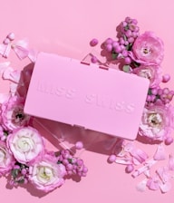 A pink rectangular box with embossed lettering surrounded by various pink and white flowers and berries on a matching pink background.