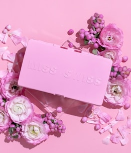 A pink rectangular box with embossed lettering surrounded by various pink and white flowers and berries on a matching pink background.